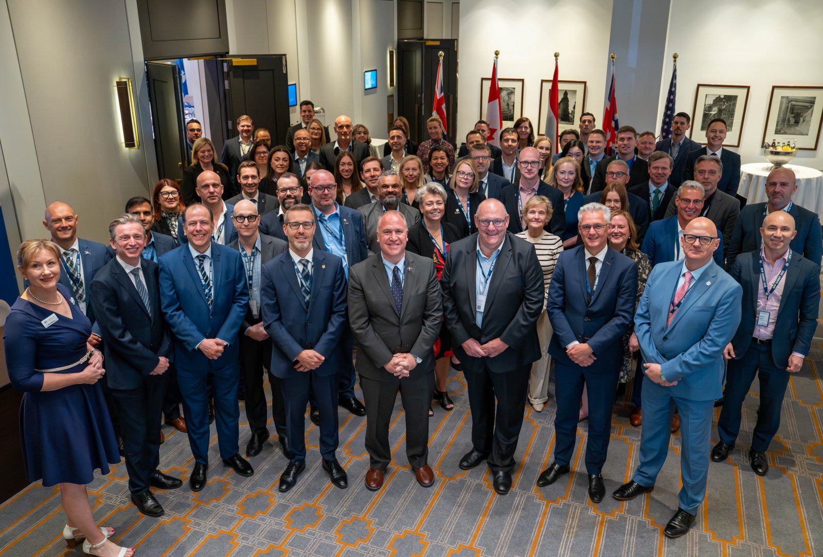 The picture shows a large group of the J5 chiefs, other delegation members and participants of the GFIP summit 2025. Its a mix of men and women. The picture is taken from above and all the people in the picture are looking up to the camera. In the front row are the 5 J5 chiefs.