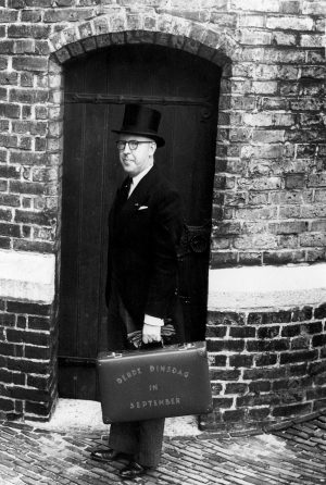 Zwartwit foto van minister van financiën in 1945, Piet Lieftinck. Hij staat afgebeeld door een houten deur in een zwarte knielange jas, een hoge zwarte hoed, en een bril. In zijn linkerhand heeft hij het koffertje met de tekst Derde DInsdag in September.