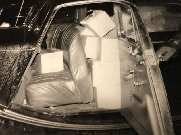 Een zwart wit fot van een donkere auto met openstaand portier, waarvan de hele achterbank is volgepropt met grote witte blokken boter.