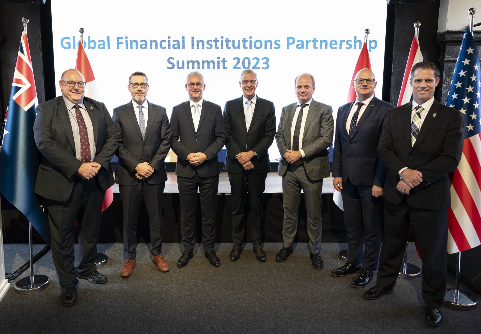 Groepsfoto op een podium met op het scherm de tekst Global Financial Institutions Partnership, daarvoor een groepsfoto van de 5 J5 chiefs, staatssecretaris van Rij en DG Belastingdienst Peter Smink. 
